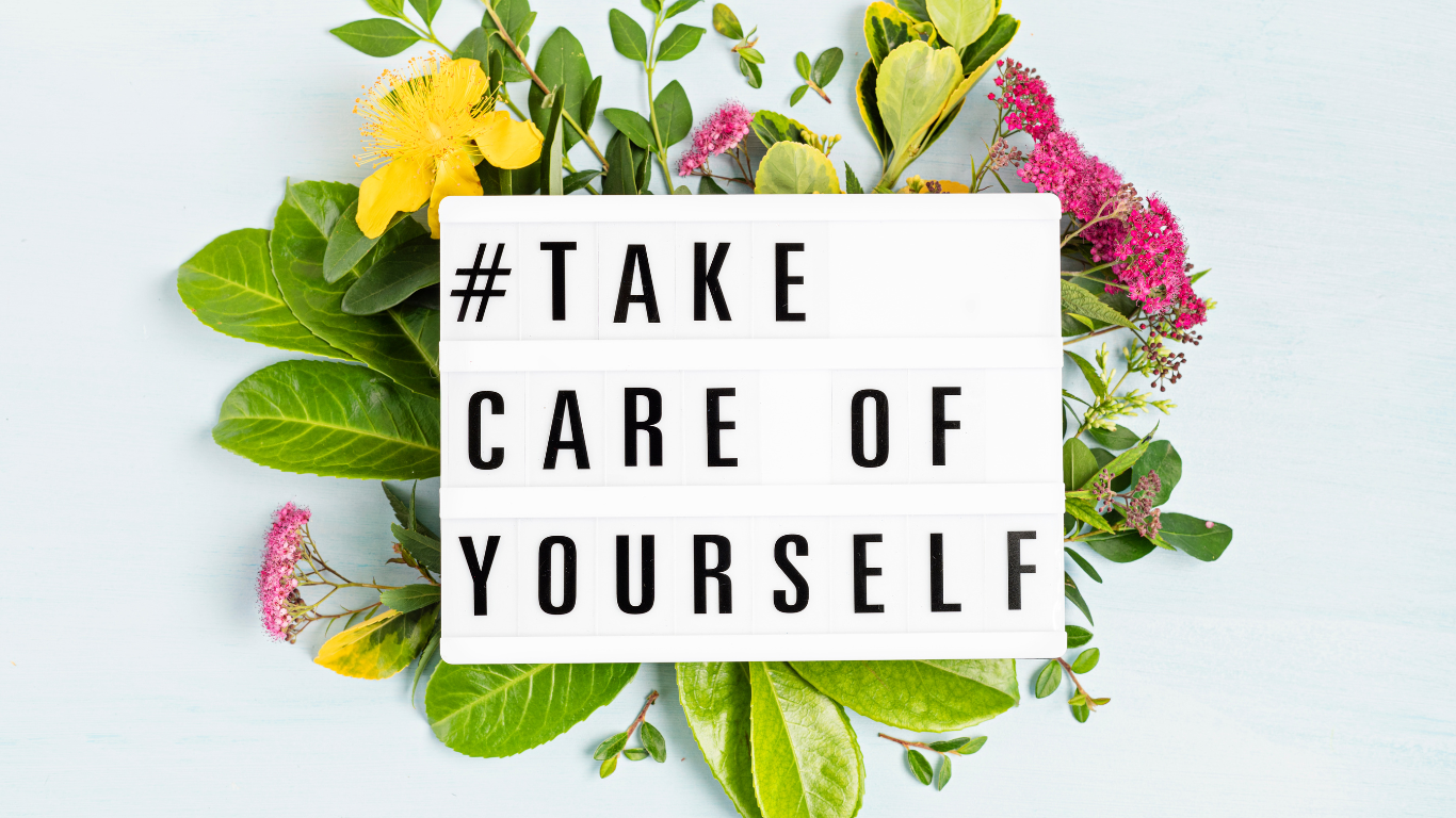 A sign that says Take Care of Yourself surrounded by flowers and greenery